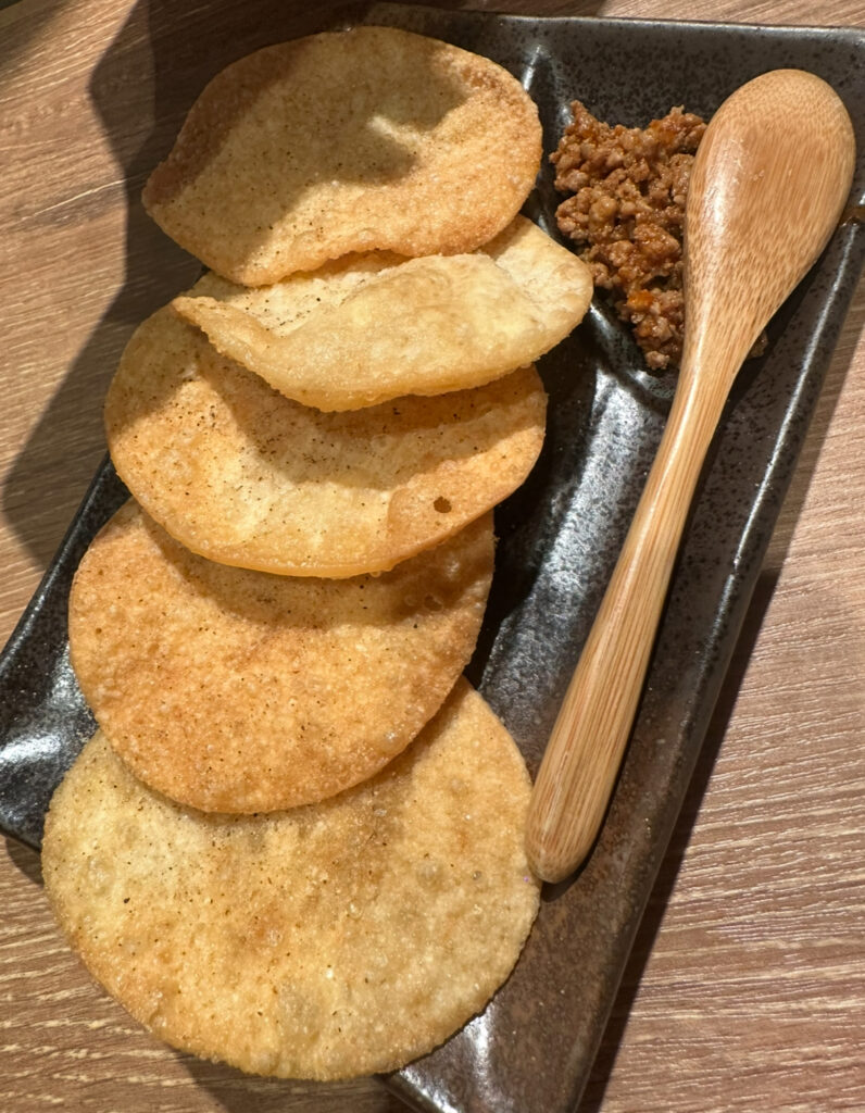 黒い長方形の皿に盛り付けられたパリパリに揚げた餃子の皮せんべい5枚と、横に添えられた肉味噌と木製スプーン