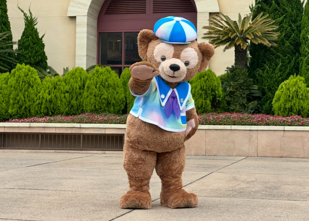 東京ディズニーシーのエントランス付近で、カラフルな衣装を着て手を振るダッフィー。ダッフィー&フレンズのごあいさつ(ステージグリ)