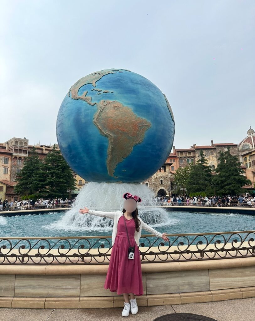 東京ディズニーシーのアクアスフィアを背景に、ピンクのワンピースとミニーのカチューシャ姿でポーズをとるブログ運営者のCOCO。