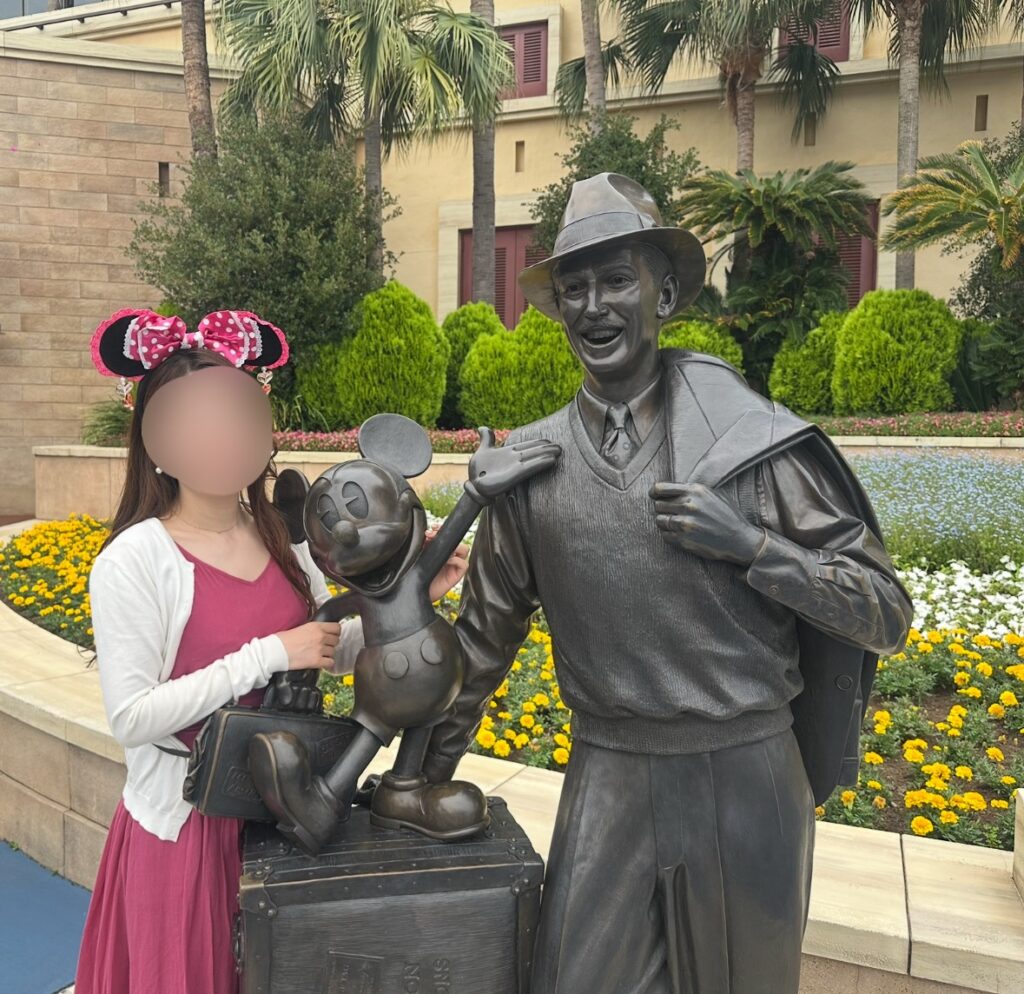 ディズニーシーのエントランス付近にある「ウォルト・ディズニーとミッキー」のブロンズ像と記念写真を撮るブログ運営者COCO。色とりどりの花壇と南国風の木々に囲まれたフォトスポット。