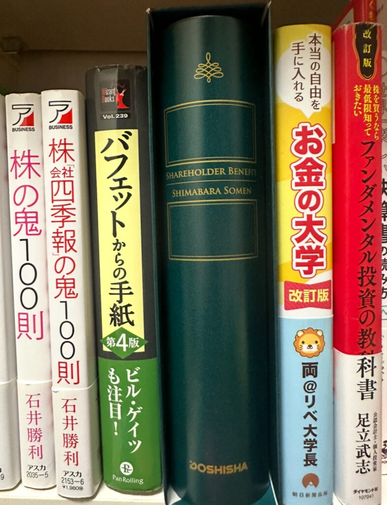 本棚に並ぶ投資や経済関連の書籍の中に、ドウシシャの株主優待「SHAREHOLDER BENEFIT SHIMABARA SOMEN」の緑色のギフトボックスが本のように収納されている様子。右には『お金の大学』や『ファンダメンタル投資の教科書』などの書籍も並んでいる。