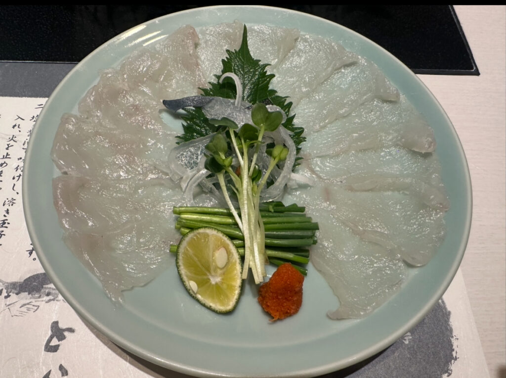 薄造りされたてっさ(ふぐ刺し)が美しく円形に盛りつけられた大皿。中央には薬味として大葉や小ねぎ、もみじおろし、すだちが添えられている。