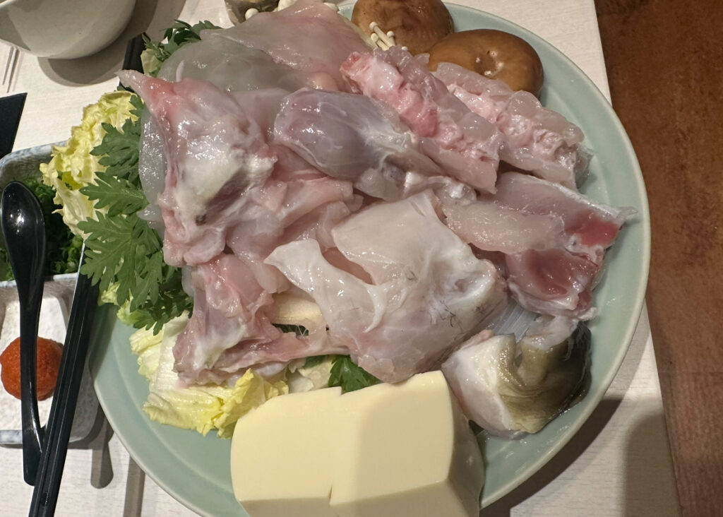 ふぐ鍋用の大皿に、ぶつ切りのふぐ身や皮、豆腐、白菜、春菊、椎茸などの鍋具材がたっぷりと盛られている。