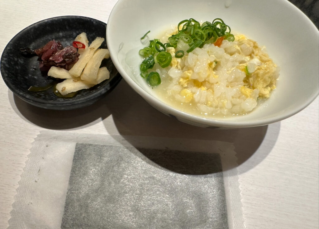 卵とネギを加えて仕上げたふぐ雑炊が白い器に盛られ、黒い小皿にたくあんや梅風味の漬物が添えられている。