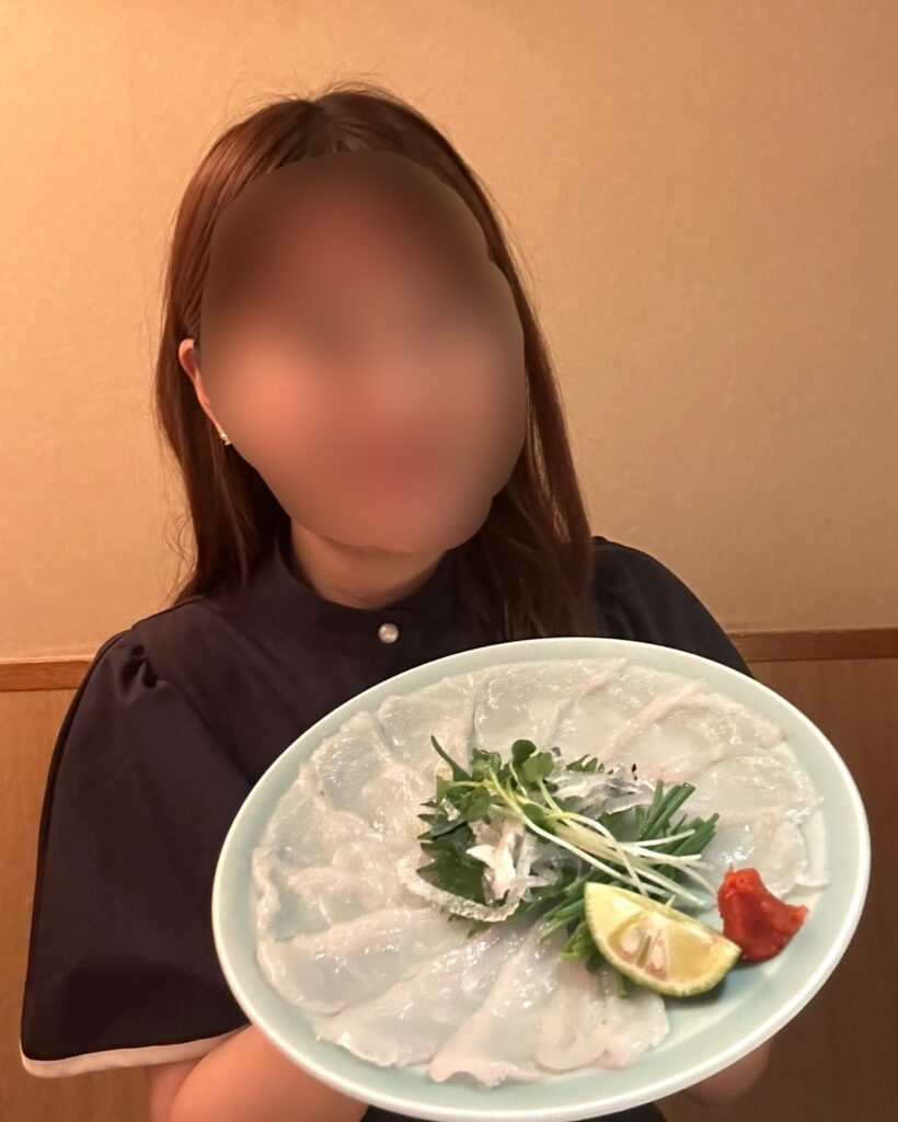 ブログ運営者COCOが大皿に盛りつけられたふぐ刺し(てっさ)を手に持ち、微笑む様子。※顔はぼかしを入れて掲載しています。
