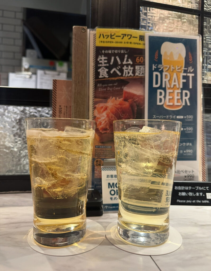 恵比寿ハイボールバー店内のカウンター席で撮影された写真。2杯のハイボールグラスがテーブルに置かれ、背景には「生ハム食べ放題」や「ドラフトビール」のポスターが並んでいる。カジュアルな居酒屋の雰囲気が伝わる一枚。