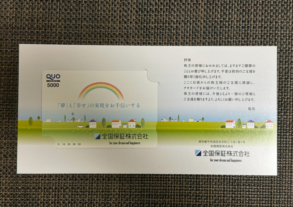 全国保証の株主優待として届いた5,000円分のQUOカードとメッセージ付き台紙。虹と街並みが描かれた優しいデザインで、『夢と幸せの実現をお手伝いする』というメッセージが印字されている。