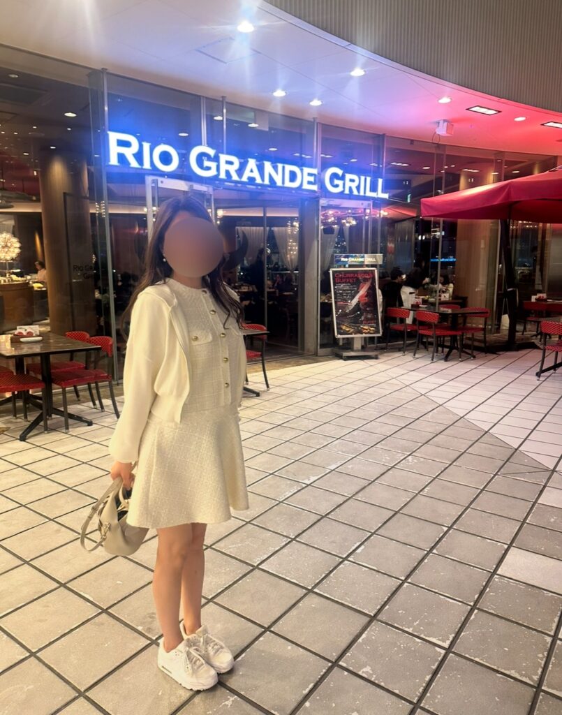 RIO GRANDE GRILL横浜ベイクォーター店の入口前で撮影したブログ運営者COCO（※顔はぼかしを入れて掲載しています）。店内はガラス張りで明るく、赤いパラソル付きテラス席も見える。