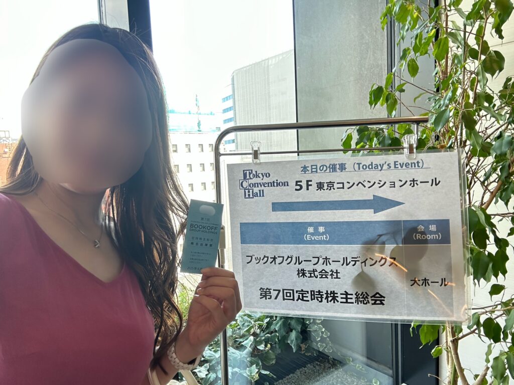 ブログ運営者COCOが、BOOKOFF第7回定時株主総会の出席表を手にし、会場案内板の前で撮影している様子。※顔はぼかしを入れて掲載しています。