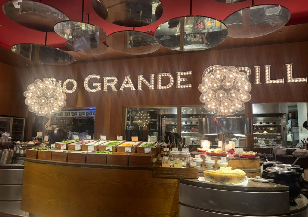RIO GRANDE GRILL横浜ベイクォーター店のサラダバーエリア。色とりどりの野菜や前菜が並び、奥にはオープンキッチンとブラジル国旗の装飾が見える。上部には煌びやかなシャンデリアが輝く。
