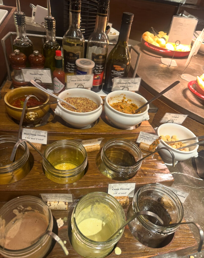 RIO GRANDE GRILL横浜ベイクォーター店のサラダバーに並ぶ各種ドレッシングと調味料。シーザードレッシング、サウザンアイランド、ブラッドオレンジ、バルサミコ酢、オリーブオイル、粒マスタードなどが並ぶ。