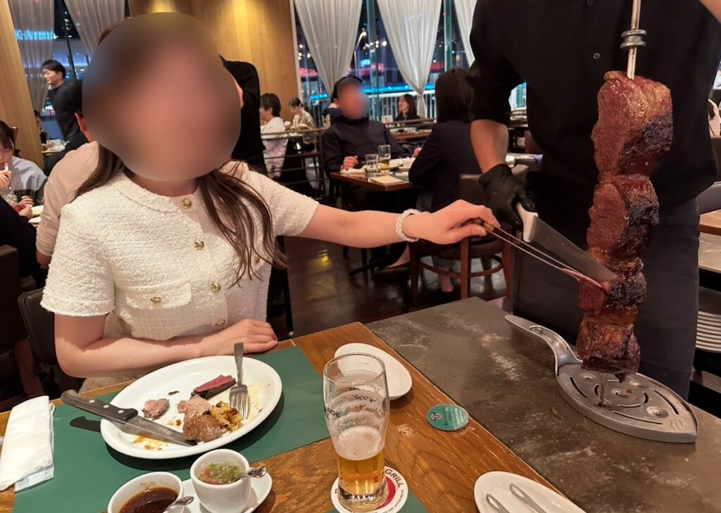 RIO GRANDE GRILL横浜ベイクォーター店でシュラスコを体験するブログ運営者COCO（※顔はぼかしを入れて掲載しています）。テーブルでスタッフが串からピッカーニャ（イチボ）を切り分ける様子。手前にはソースやビールが並ぶ。