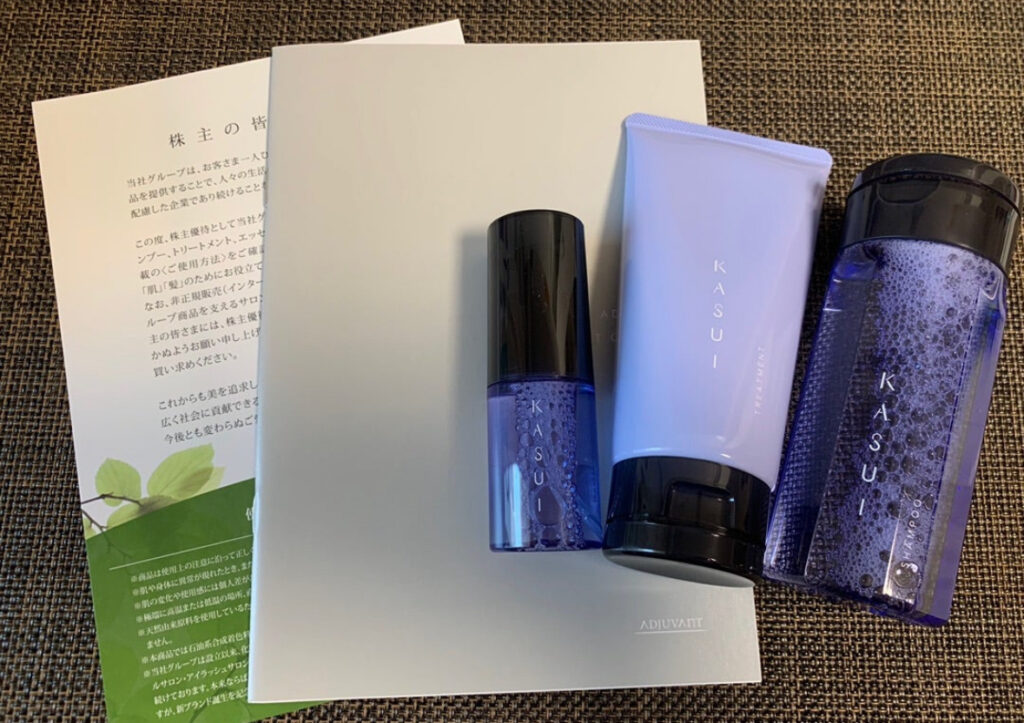 2022年に届いたアジュバン株主優待品「KASUI」シリーズ。シャンプー・トリートメント・エッセンスの3点セットと、株主向け案内冊子が並んでいる。紫と黒を基調とした高級感あるパッケージデザインが特徴。