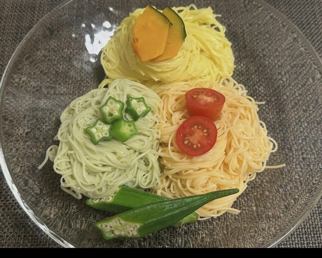 「エスリードの株主優待でもらった『夏野菜を使った三輪素麺』をガラス皿に盛り付けた写真。トマト素麺にはミニトマト、かぼちゃ素麺には蒸しかぼちゃ、オクラ素麺には輪切りオクラをトッピングし、見た目にも華やかな夏らしい一皿に仕上がっている。」