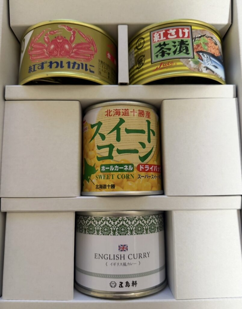alt="ホッカンホールディングスの株主優待2025で届いた食品缶詰セット。紅ずわいがに缶、紅さけ茶漬け缶、スイートコーンドライパック、五島軒のイギリス風カレー缶が並ぶ。"
