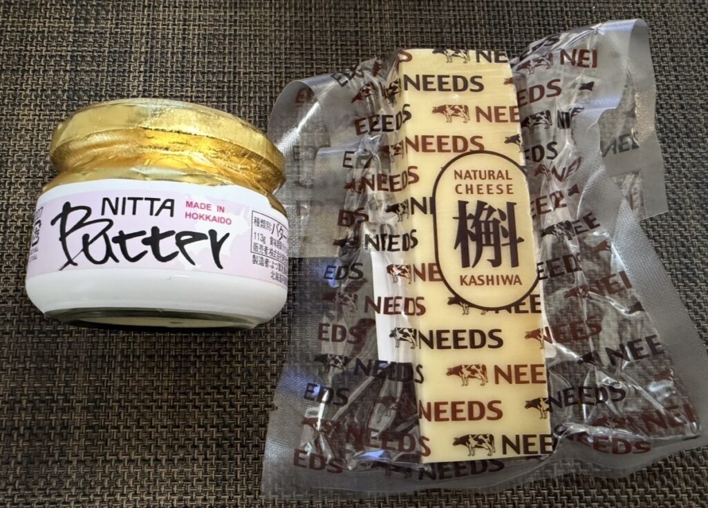 ニッタ（5186）の株主優待として届いたバターとチーズのセット。左側にゴールドのフタの北海道バター、右側に「NEEDS」ブランドのナチュラルチーズ「柏」が並んでいる。
