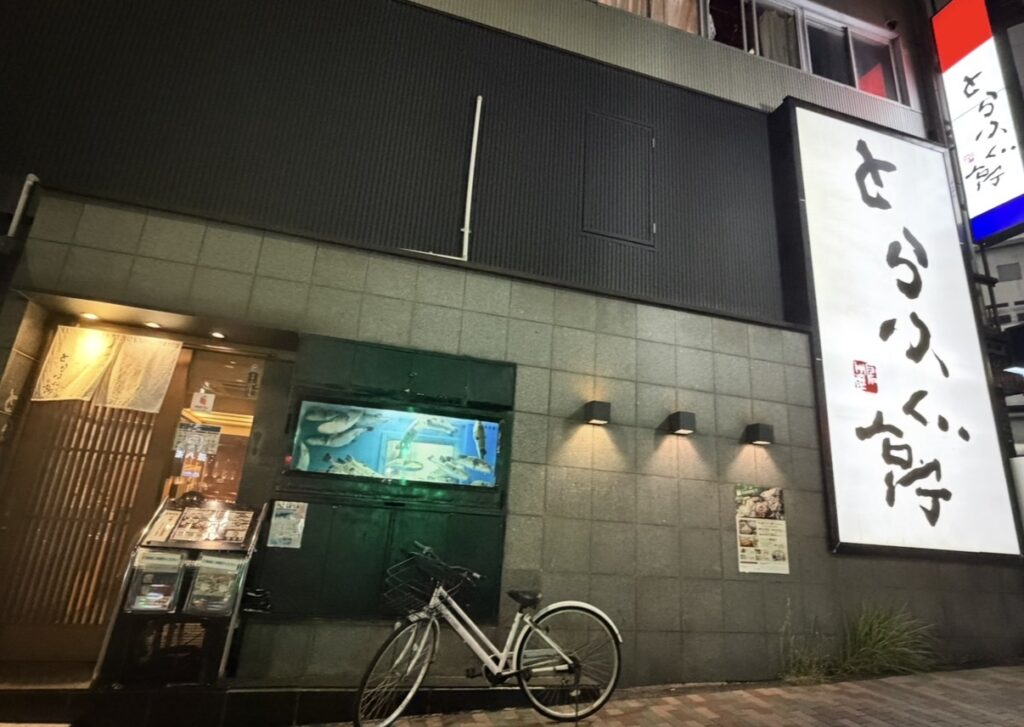 とらふぐ亭 目黒店の外観。大きな白い看板と、水槽に泳ぐふぐが印象的な店舗正面。夜のライトアップで落ち着いた雰囲気が漂う。
