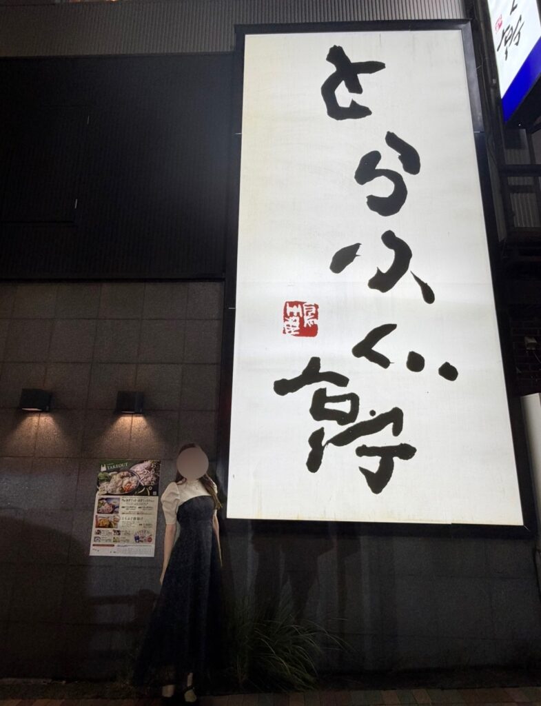 とらふぐ亭 目黒店の大きな看板の前で撮影された、ブログ運営者COCOの写真。和風の外壁と照明に包まれた店構えの前で、上品なドレス姿のCOCOが立っている様子。※顔はぼかしを入れて掲載しています。