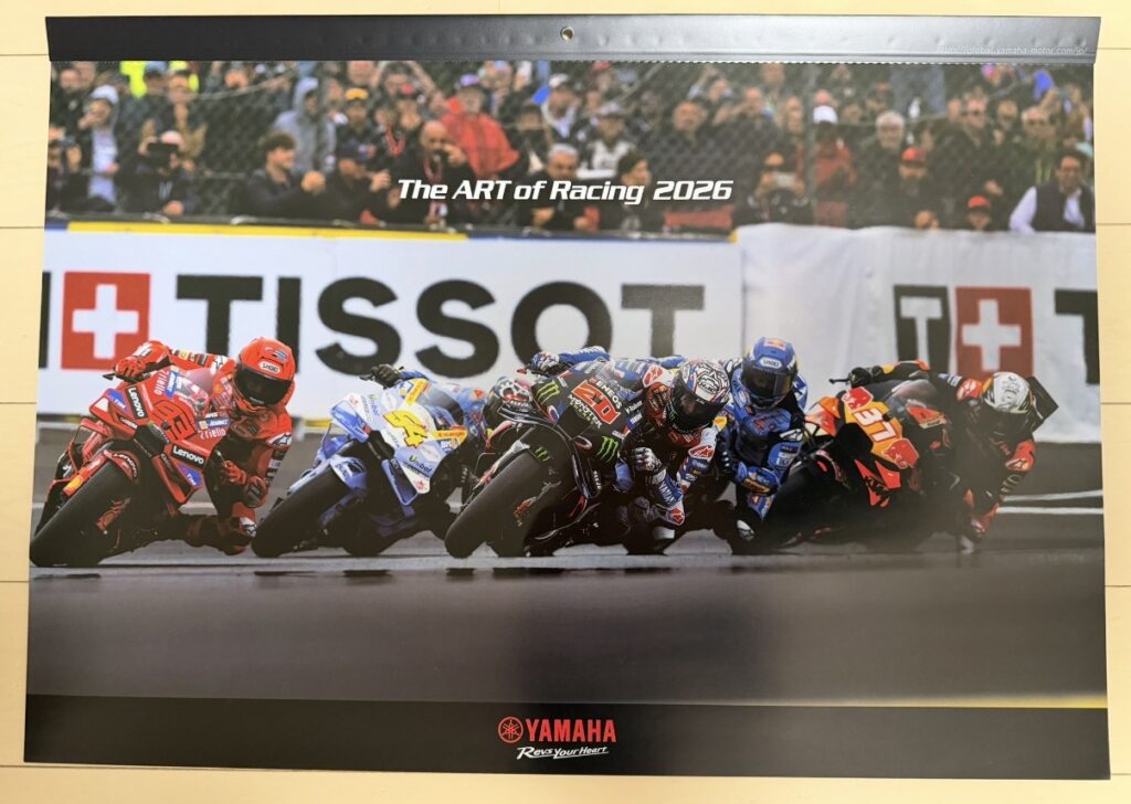 ヤマハ発動機のレーシングカレンダー2026年版の表紙写真。『The ART of Racing 2026』の文字が中央上部に配置され、MotoGP World Championshipでファビオ・クアルタラロ(Fabio Quartararo)がYZR-M1に乗って激しいレースバトルを繰り広げるシーンが写されている。下部にはYAMAHA Rev Your Heartのロゴが入ったデザイン。