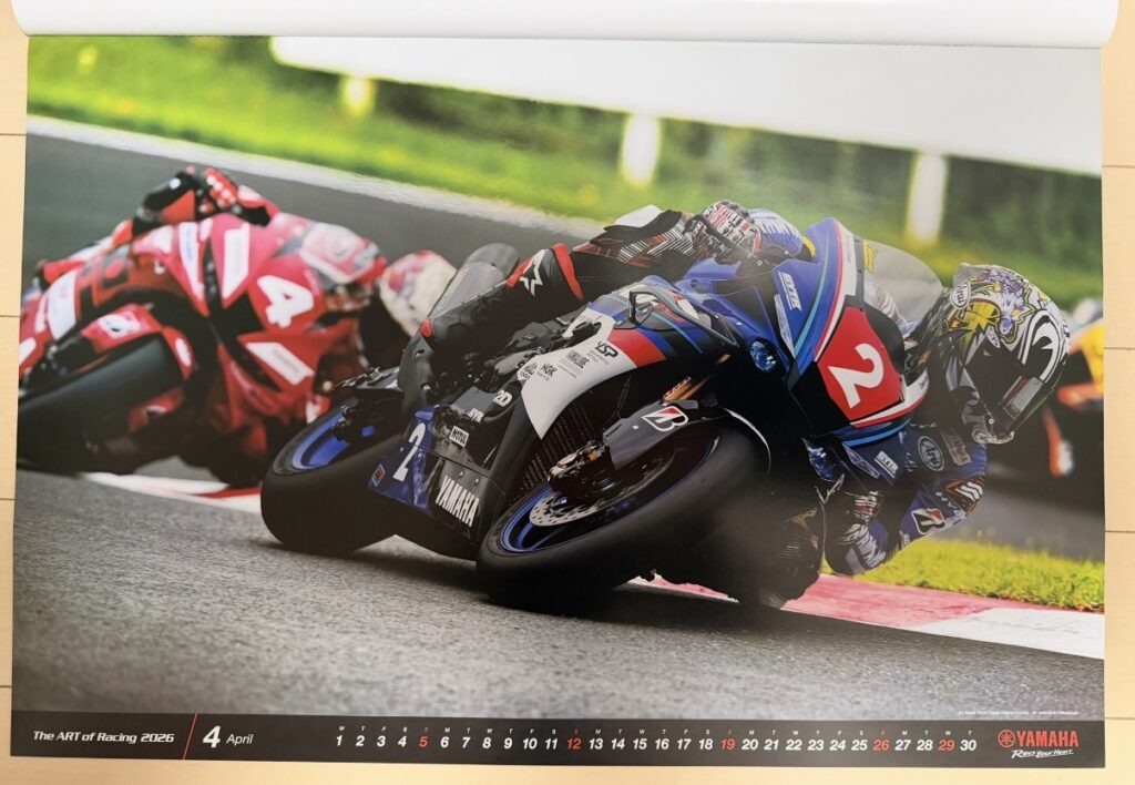 ヤマハ発動機のレーシングカレンダー2026年版・4月の写真。全日本ロードレース選手権(All Japan Road Race Championship)SUGOのスーパーバイクレースで、中須賀克行(Katsuyuki Nakasuga)がYZF-R1に乗り、深いバンク角でコーナーを攻めるシーンが写されている。背後には他のライダーも続き、日本国内レース特有の迫力と緊張感が漂う一枚。