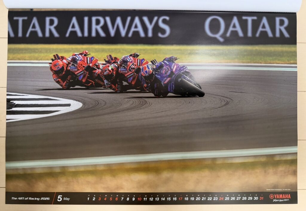 ヤマハ発動機のレーシングカレンダー2026年版・5月の写真。MotoGP World ChampionshipイギリスGPで、ジャック・ミラー(Jack Miller)がYZR-M1を鋭くバンクさせながらコーナーを攻め、背後に複数のライダーが続く緊迫したバトルシーンが写されている。路面には黒いラインが残り、MotoGP特有のスピード感が伝わる一枚。