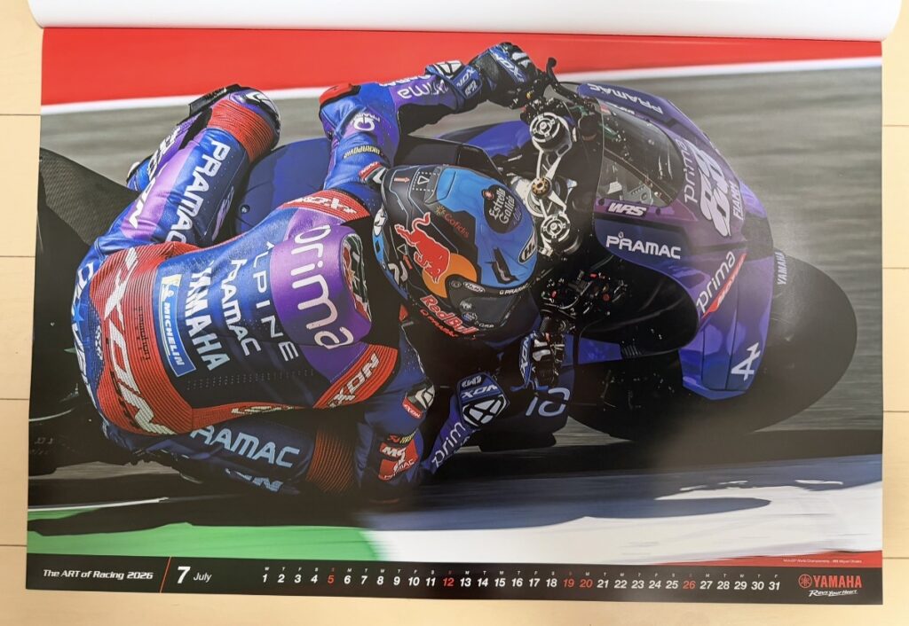 ヤマハ発動機のレーシングカレンダー2026年版・7月の写真。MotoGP世界選手権イタリアGPで、ミゲル・オリベイラ(Miguel Oliveira)がYZR-M1を極限までバンクさせながらコーナーを攻める姿が写されている。鮮やかなレッドとパープルのレーシングスーツ、マシンの曲線が美しく際立ち、MotoGP特有のスピード感と迫力が伝わる一枚。