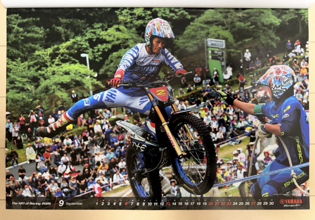 ヤマハ発動機のレーシングカレンダー2026年版・9月の写真。全日本トライアル選手権MOTEGIラウンドで、黒山健一(Kenichi Kuroyama)が電動トライアルバイク TY-E 3.0 を操り、空中でバランスを取りながら障害物を越える瞬間を捉えた一枚。観客席の前で繊細かつダイナミックな動きが際立ち、トライアル競技の高度な技術が伝わる写真。