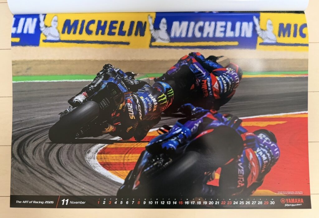 ヤマハ発動機のレーシングカレンダー2026年版・11月の写真。MotoGP世界選手権バレンシアGPで、Álex Rins、Jack Miller、Miguel Oliveira らがYZR-M1を駆り、タイトなコーナーを深いリーン角で走行する迫力のシーン。鮮やかなミシュランの看板とカラフルな路面が背景に映え、レース終盤の緊張感とスピード感が伝わる一枚。