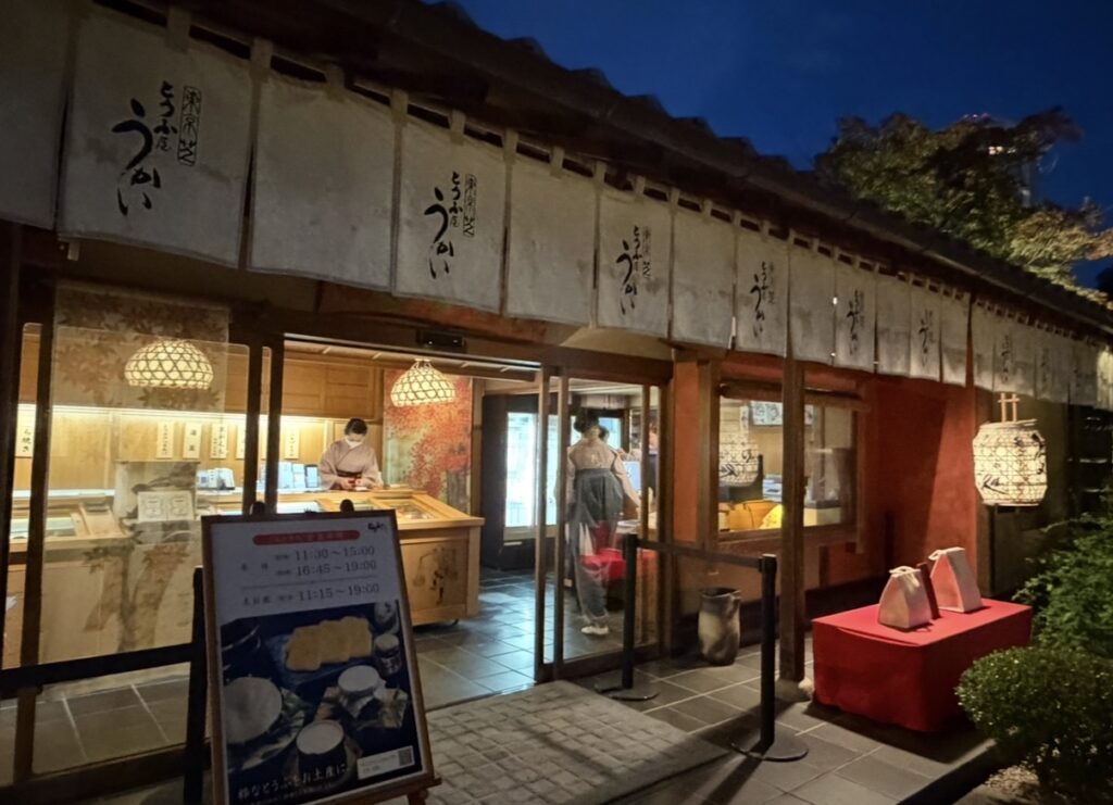 とうふ屋うかい芝店で、門をくぐって少し歩いた場所にあるお土産処の外観。暖簾がかかる落ち着いた和の建物で、店内ではスタッフが商品を整え、温かな灯りが夜の雰囲気を彩っている。