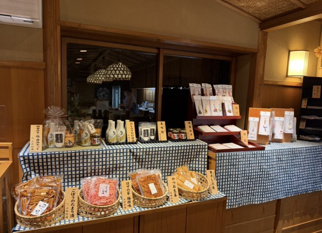 とうふ屋うかい芝店のお土産処の店内で、調味料やお菓子、季節の小物が整然と並んだディスプレイ。和の温かみを感じる木の棚や柔らかな照明が、落ち着いた雰囲気を演出している。