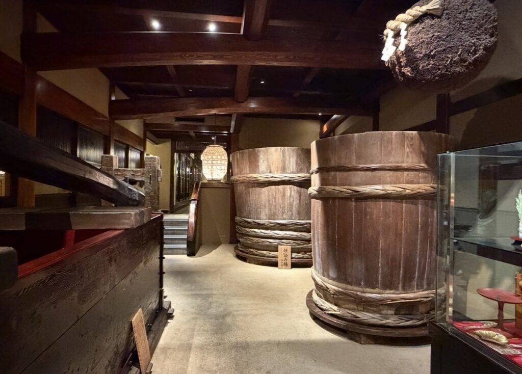とうふ屋うかい芝店の通路に展示された古い味噌樽や日本酒の仕込み道具。木の梁や土壁が残る趣ある空間で、かつての造り酒屋の雰囲気を感じられる展示の写真。