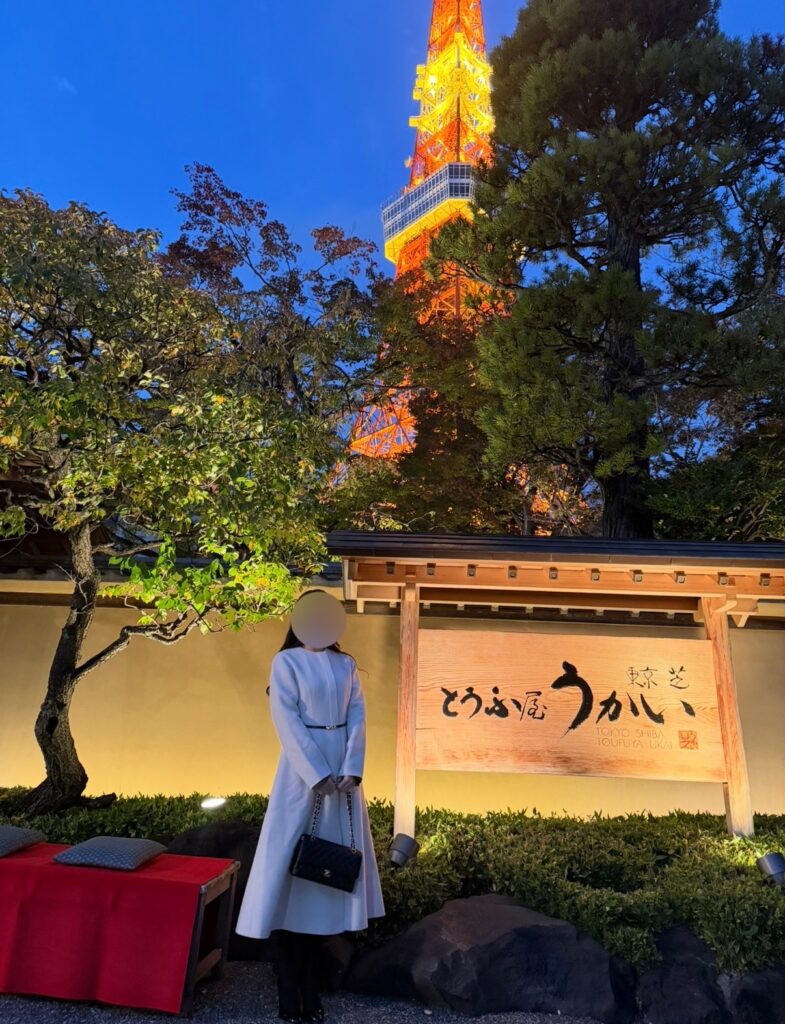 ライトアップされた東京タワーを背景に、とうふ屋うかい芝店の入口前で立つブログ運営者COCOの写真。白いロングコートを着て、和の門構えと庭木、看板が美しく照らされた夜の雰囲気を写した一枚。※顔はぼかしを入れて掲載しています。