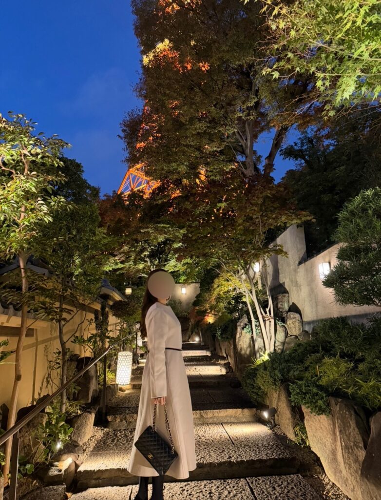 とうふ屋うかい芝店の門をくぐった直後の石畳の小道で、ライトアップされた東京タワーを見上げるブログ運営者COCOの写真。白いロングコート姿で、柔らかな灯りに照らされた庭木や石垣が、和の雰囲気と非日常感を演出している一枚。※顔はぼかしを入れて掲載しています。