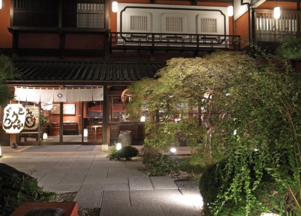 とうふ屋うかい芝店の夜の入口の様子。白い暖簾がかかったお迎え処が柔らかな灯りに照らされ、庭木と石畳が落ち着いた和の雰囲気を演出している。静かで上品な空気が伝わる玄関の写真。