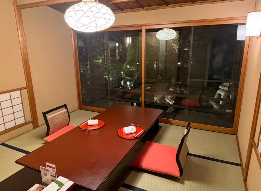 とうふ屋うかい芝店の個室。畳敷きの広い和室に赤い座布団と大きな座卓が配置され、窓の外にはライトアップされた庭園が見える落ち着いた空間の写真。
