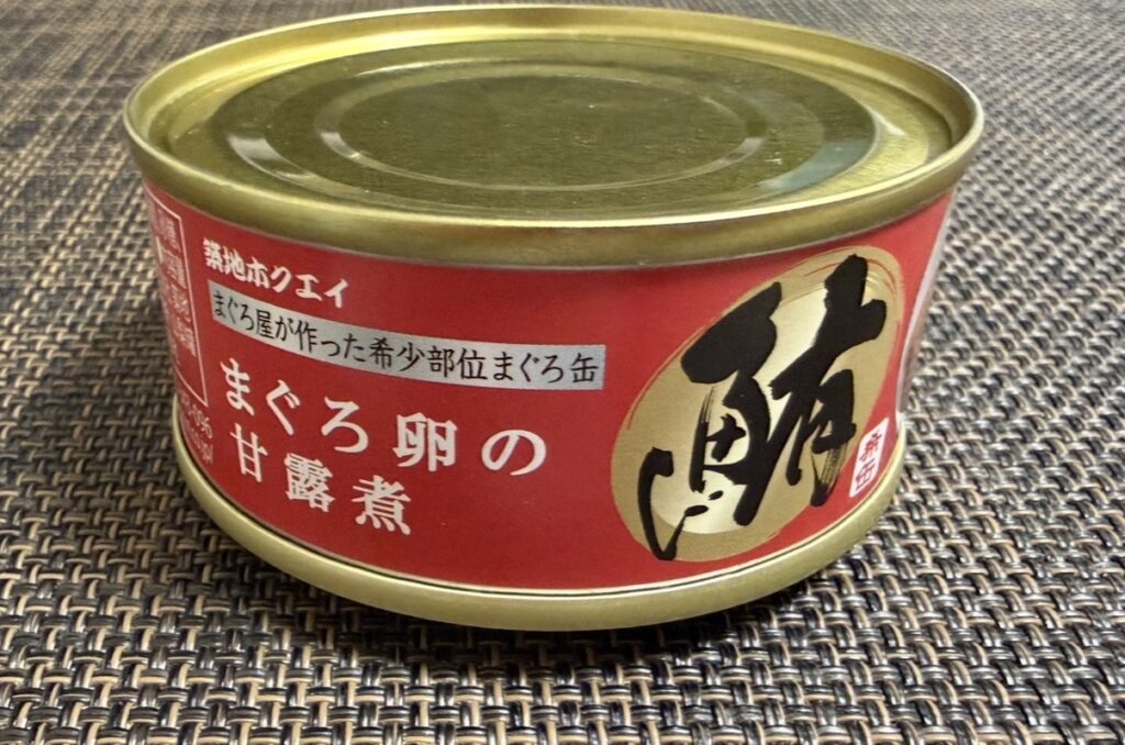 ゆうちょ銀行の株主優待で届いた築地魚河岸「希缶」シリーズ、まぐろ卵の甘露煮の缶詰。赤いラベルに大きく『鮪』の文字が入った高級感あるデザインで、テーブルの上に置かれている様子を斜めから撮影した写真。