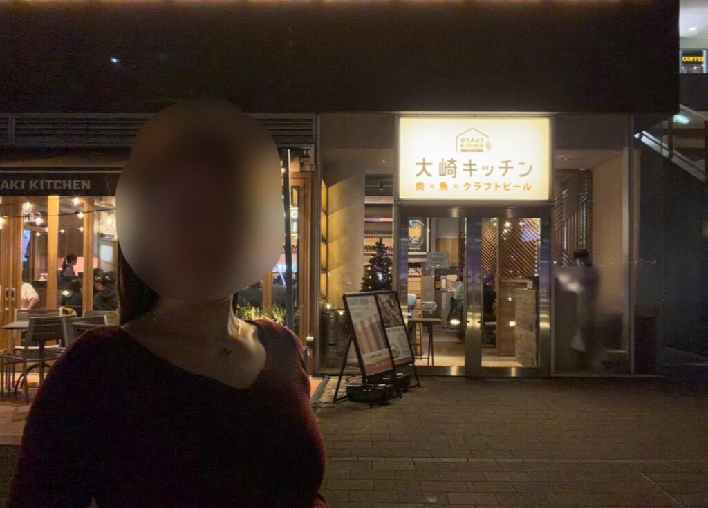 東京・大崎にある「大崎キッチン（OSAKI KITCHEN）」の店舗入口の外観。夜の時間帯に撮影された写真で、店名の看板とガラス張りの入り口が写っている。※顔はぼかしを入れて掲載しています。