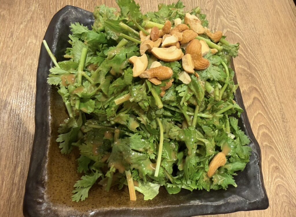 ダンダダン名物『悪魔的に旨い香菜サラダ』を盛りつけた一皿。山盛りのパクチーの上にカシューナッツがたっぷり乗り、特製ドレッシングがかかった鮮やかなサラダ。木目テーブルの上に置かれた、ボリューム感と新鮮さが伝わる写真。