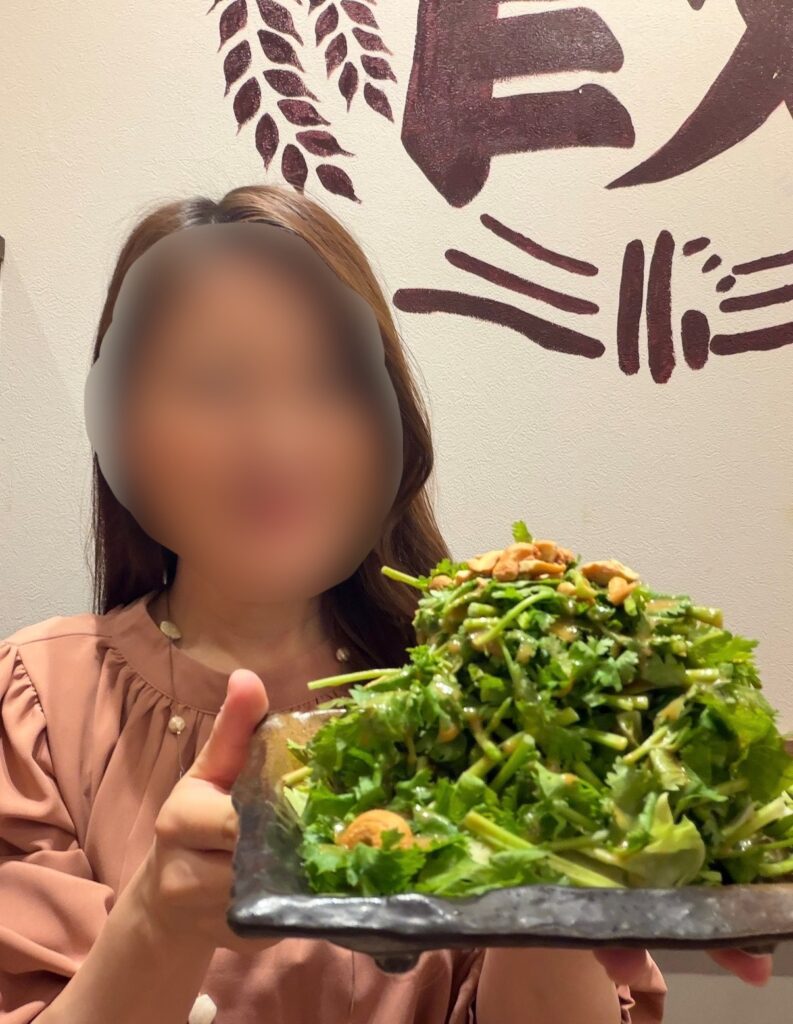 ブログ運営者COCOが『悪魔的に旨い香菜サラダ』を手に持っている様子。顔はぼかしを入れて掲載。山盛りのパクチーとカシューナッツがのったサラダを前に出す構図で、ダンダダン店内のロゴ壁が背景に写っている。