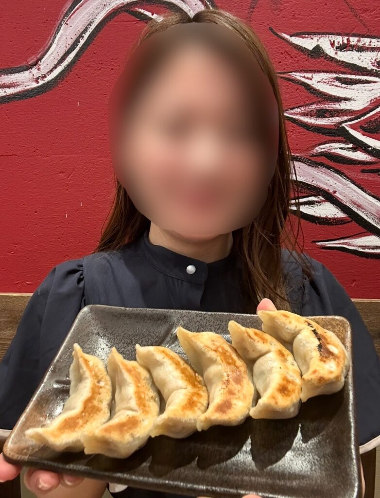 ブログ運営者COCOがダンダダン名物『肉汁焼餃子』を手に持っている様子。顔はぼかしを入れて掲載。黒い皿に並んだ餃子のもちもちした皮と焼き目がはっきり見え、赤い店内壁の前で撮影された写真。