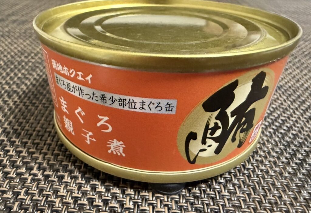 ゆうちょ銀行の株主優待カタログで選んだ築地魚河岸「希缶」シリーズのまぐろ親子煮の缶詰。オレンジ色のラベルに大きく「鮪」と書かれたデザインで、希少部位を使用した加工品を撮影した写真。
