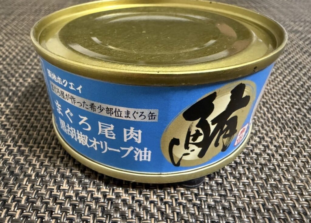 ゆうちょ銀行の株主優待で届いた築地魚河岸「希缶」シリーズのまぐろ尾肉・黒胡椒オリーブ油の缶詰。青いラベルに大きく「鮪」の文字が入ったデザインで、希少部位を使用した高級感のある缶詰をテーブルに置いて撮影した写真。
