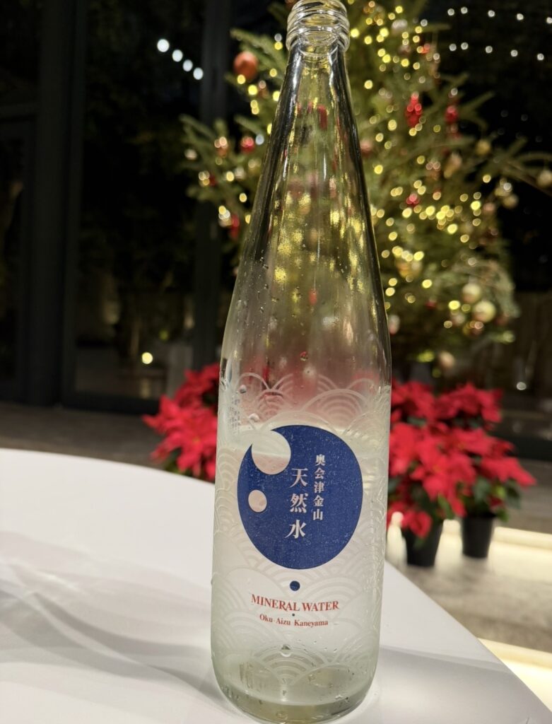 レストランで選んだガスなしのミネラルウォーター「奥会津金山 天然水」。クリスマスツリーとポインセチアを背景にしたボトルの写真