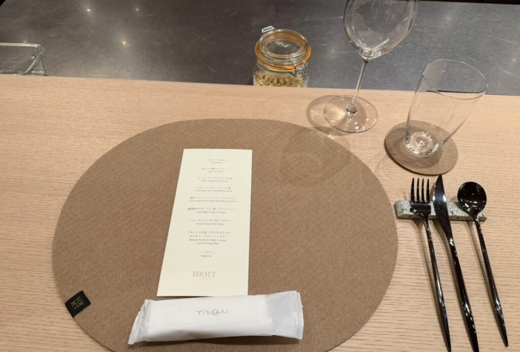 ひらまつ新店舗「HRMT STAGE」のカウンター席にセットされたカトラリーとグラス。丸いプレースマットの上にメニューが置かれたテーブルセッティングの様子。