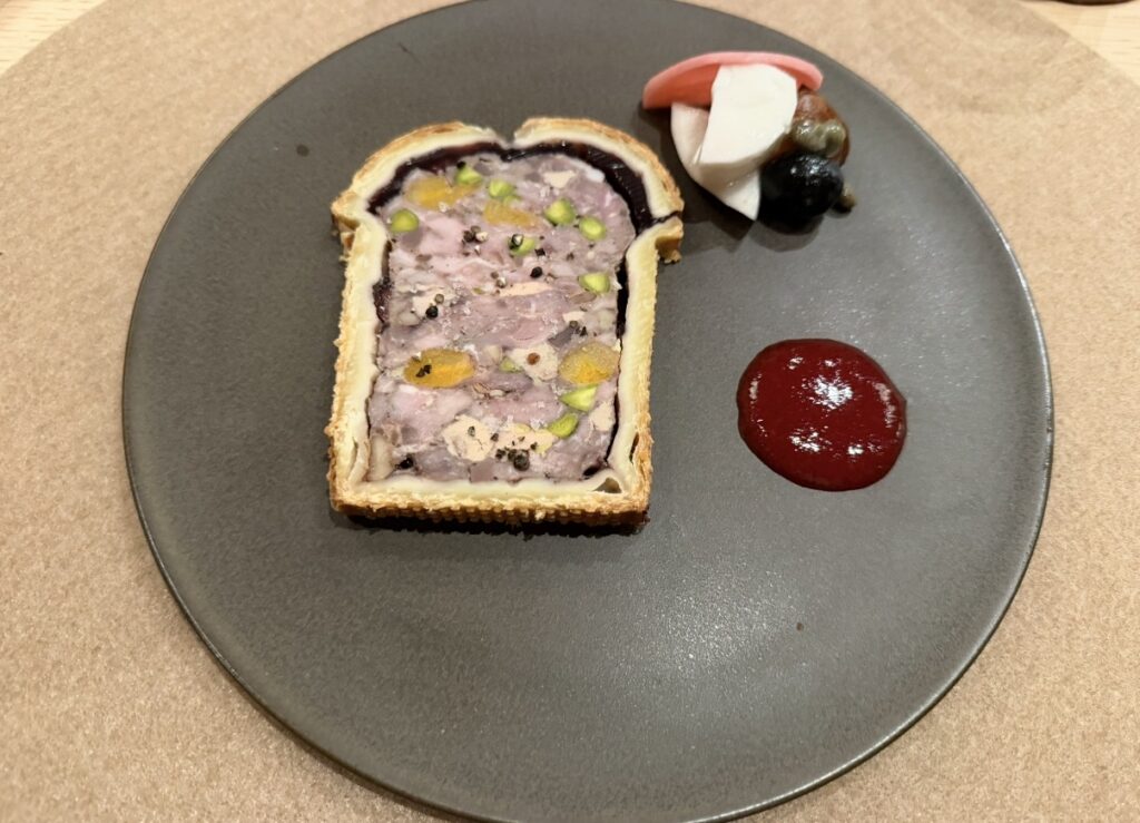 ひらまつ新店舗「HRMT STAGE」で注文した7日間かけて仕上げた自家製パテ・アンクルート。ピスタチオやドライフルーツが入った断面が美しい一皿の写真。