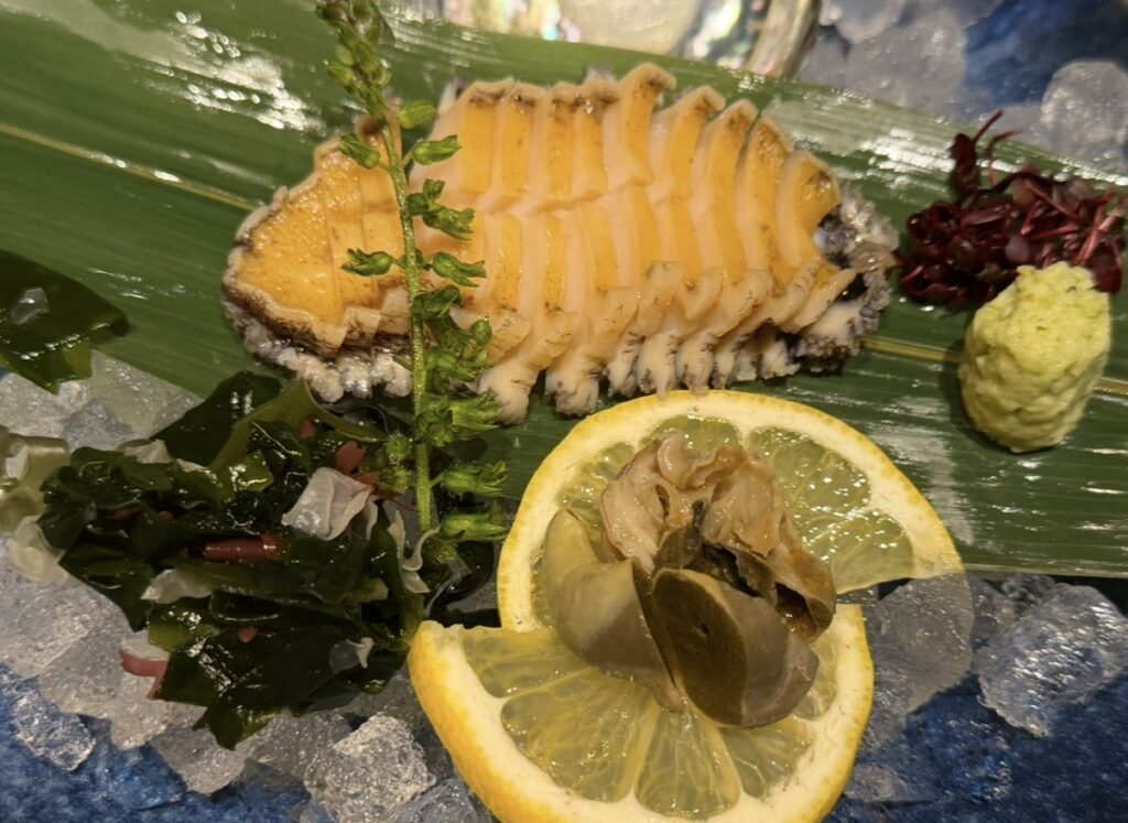 北の味紀行と地酒 北海道で注文した活蝦夷アワビ刺身。氷の上に盛り付けられた新鮮なアワビの刺身とレモン、わかめ、わさびが添えられている