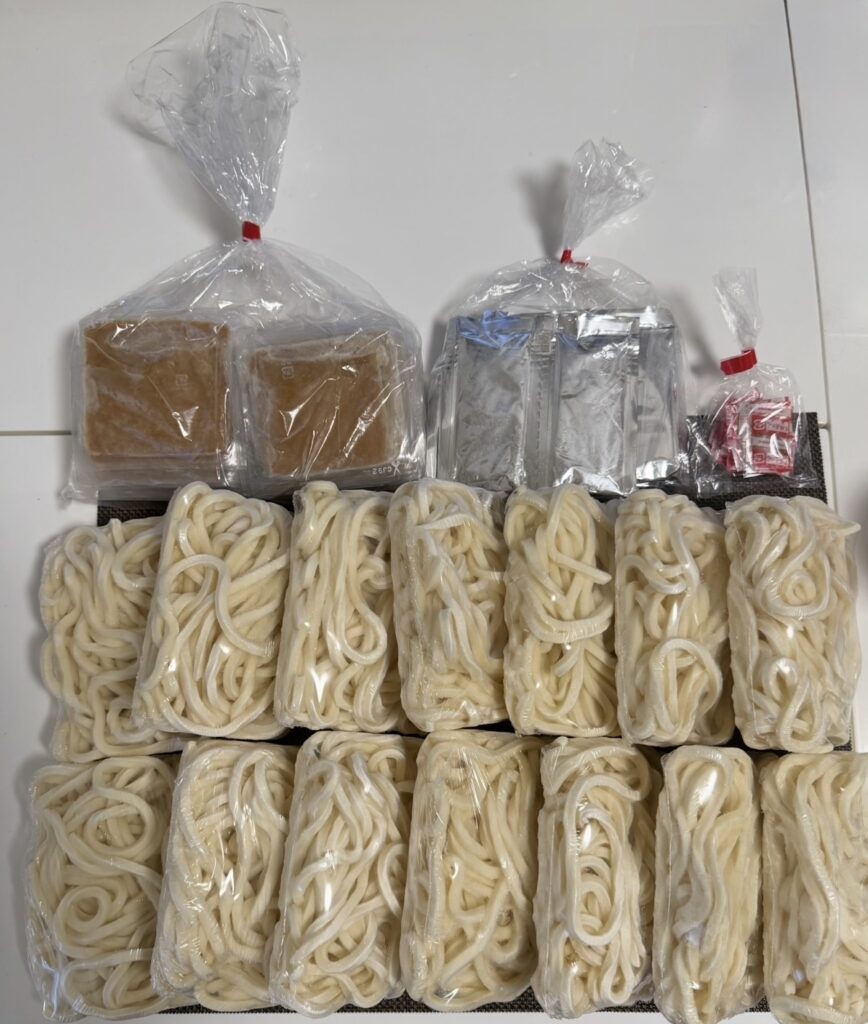 穴吹興産（8928）の株主優待で届いた冷凍さぬきうどん14食分を並べた写真。めんつゆ、味付け揚げ、七味唐辛子などの付属調味料も一緒に写っている。