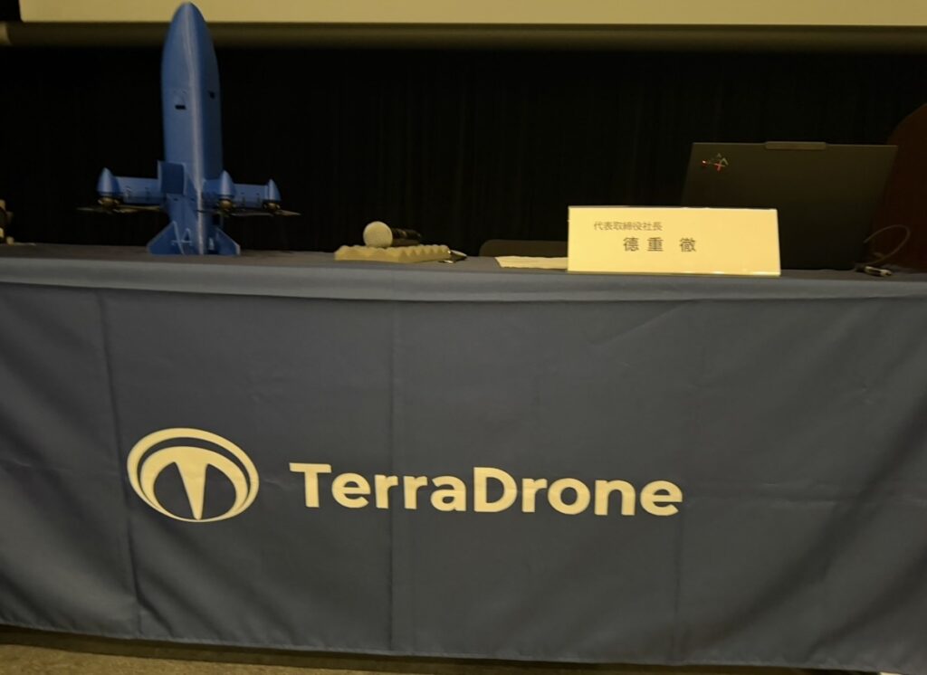 テラドローン株主総会のステージの様子。TerraDroneのロゴが入ったテーブルの上に、機体モデルやマイク、徳重徹社長のネームプレートが設置されている様子。
