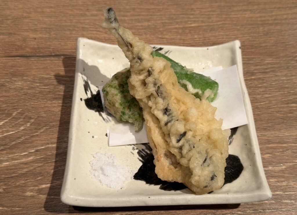 続いて登場したのは揚げ物の
**「ハタハタ天ぷらと蓮根磯辺揚げ」**です。
サクッと軽い衣で揚げられたハタハタの天ぷらは、外はカリッと中はふんわり。
焼きとはまた違った食感で、ハタハタの旨味を楽しめました。
蓮根の磯辺揚げも香ばしく、ほんのりと海苔の風味が感じられる一品。
添えられた塩でシンプルにいただくことで、素材の味をしっかり味わえました。