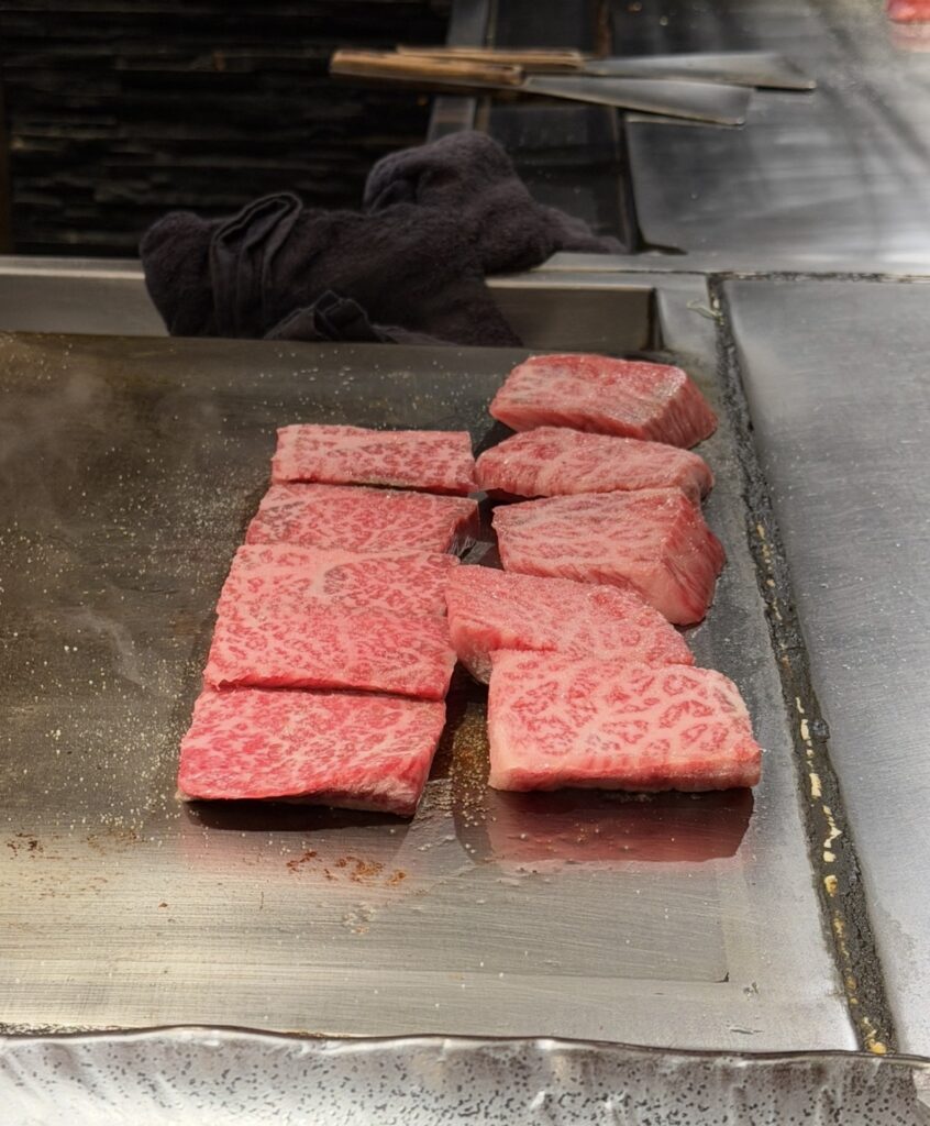 銀座のステーキ銀座本店でA5黒毛和牛ステーキが鉄板の上で焼かれている様子。美しい霜降り肉が目の前で調理されているライブ感のあるシーン